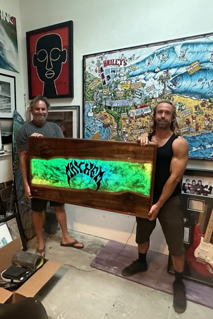 Two men holding a glowing framed artwork with a tree design indoors.